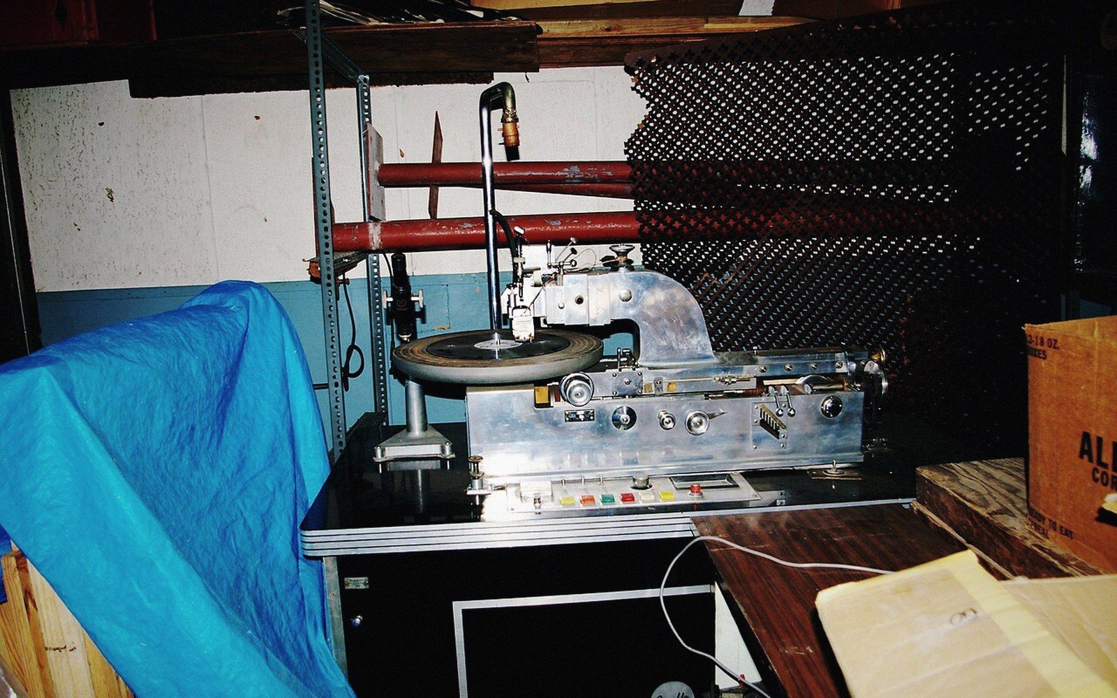 Close-up of a vinyl cutting lathe in a pressing plant, the stylus above a lacquer disc, metal surfaces catching overhead light